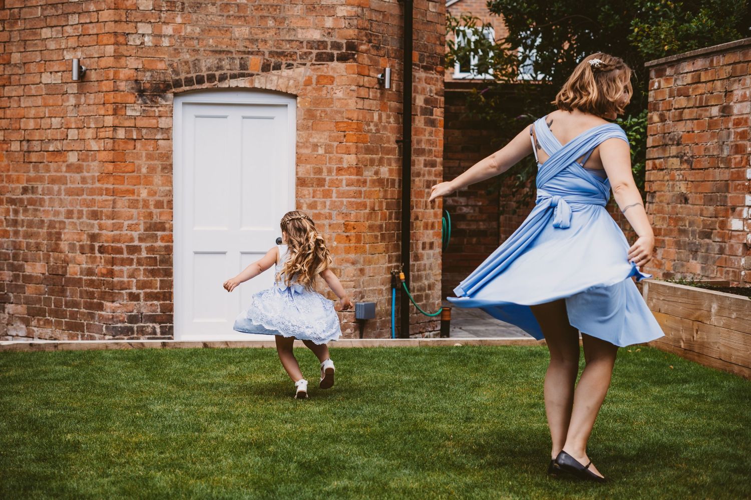 Two people in matching blue dresses twirling on a lawn beside a brick building.
