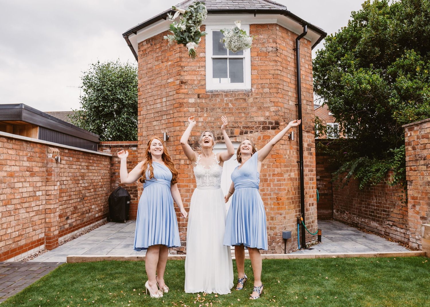 Three people pose joyfully in front of a brick building with their arms raised on a garden lawn.