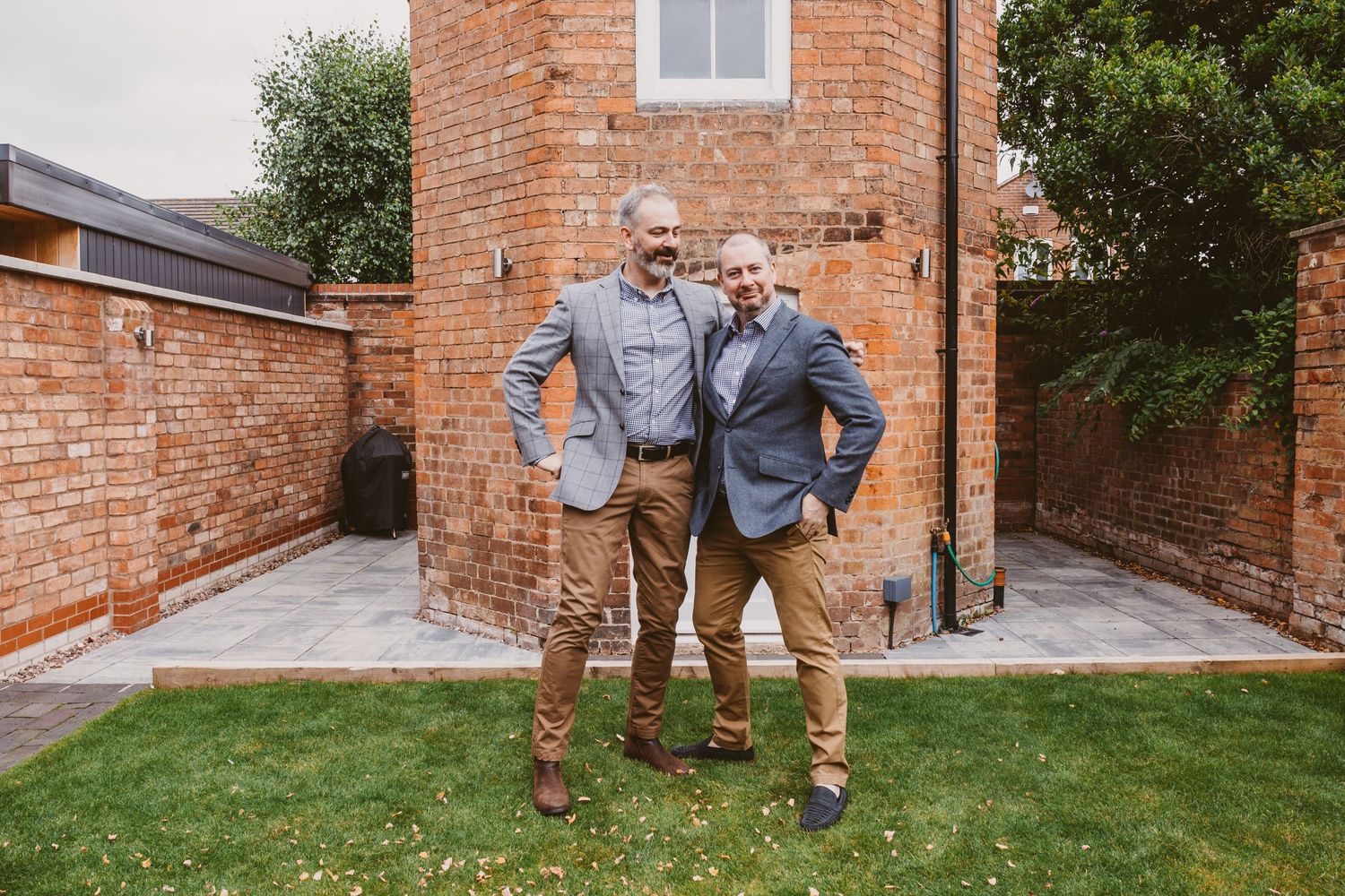 Two people wearing casual attire stand together in front of a rustic brick building with a lawn.
