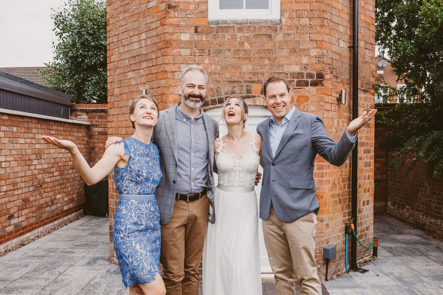 Wedding party poses in front of red brick building with garden backdrop.