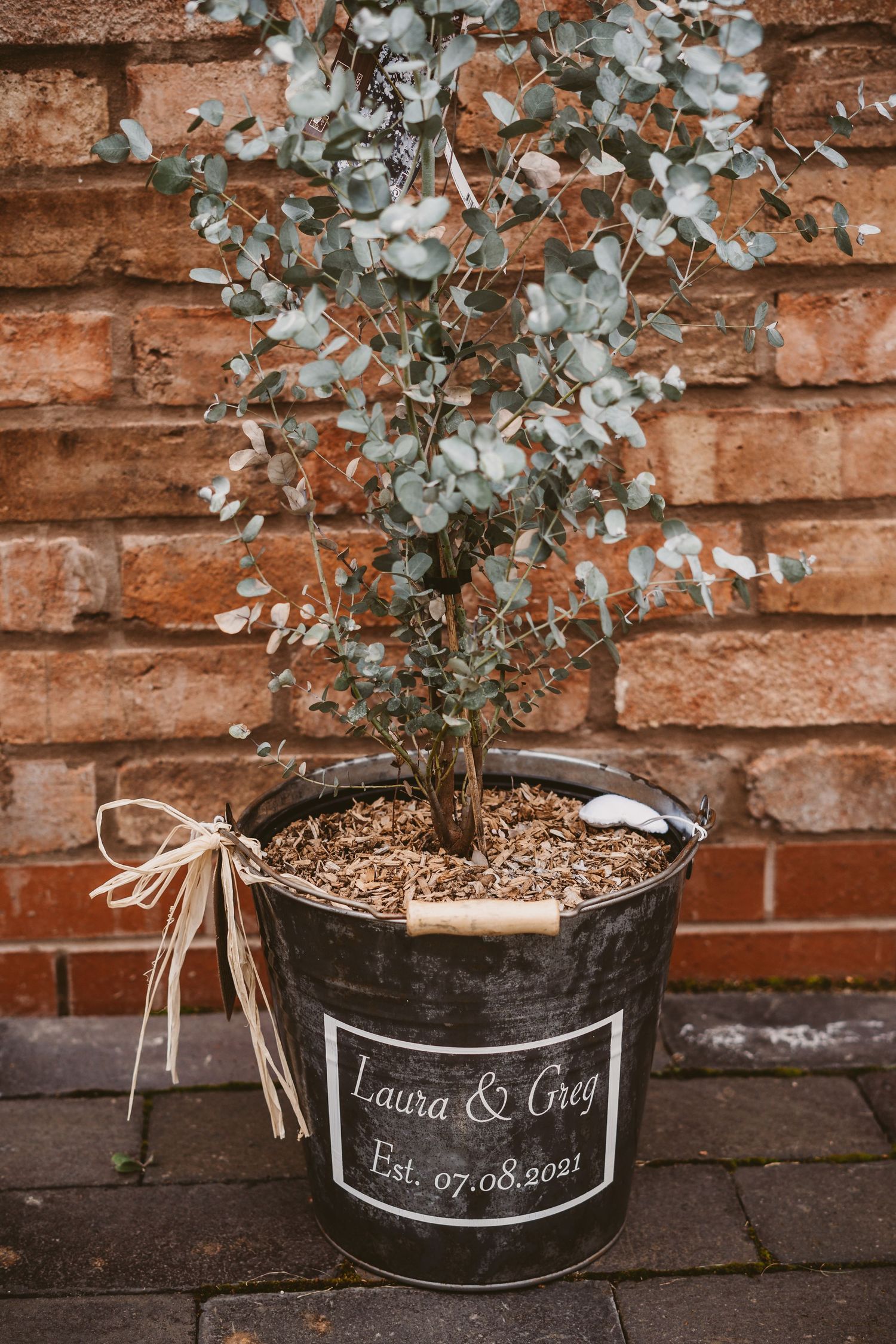 A potted eucalyptus plant in a rustic bucket with a personalized chalkboard label displaying names and date.