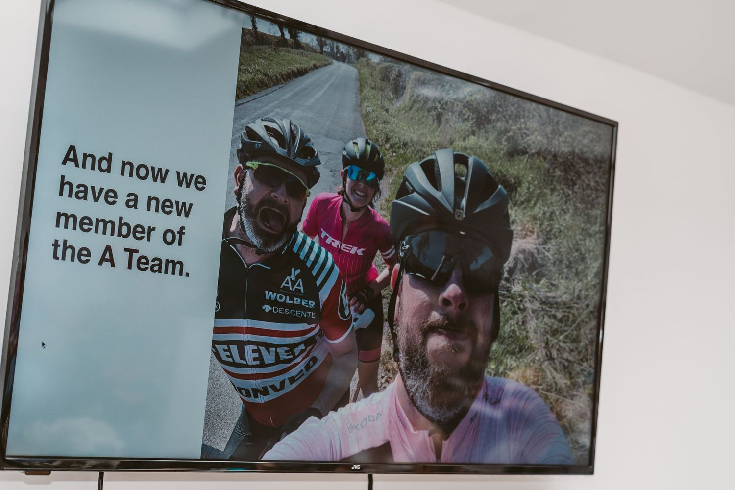 Television screen displays text about A-Team member with cyclists in background.