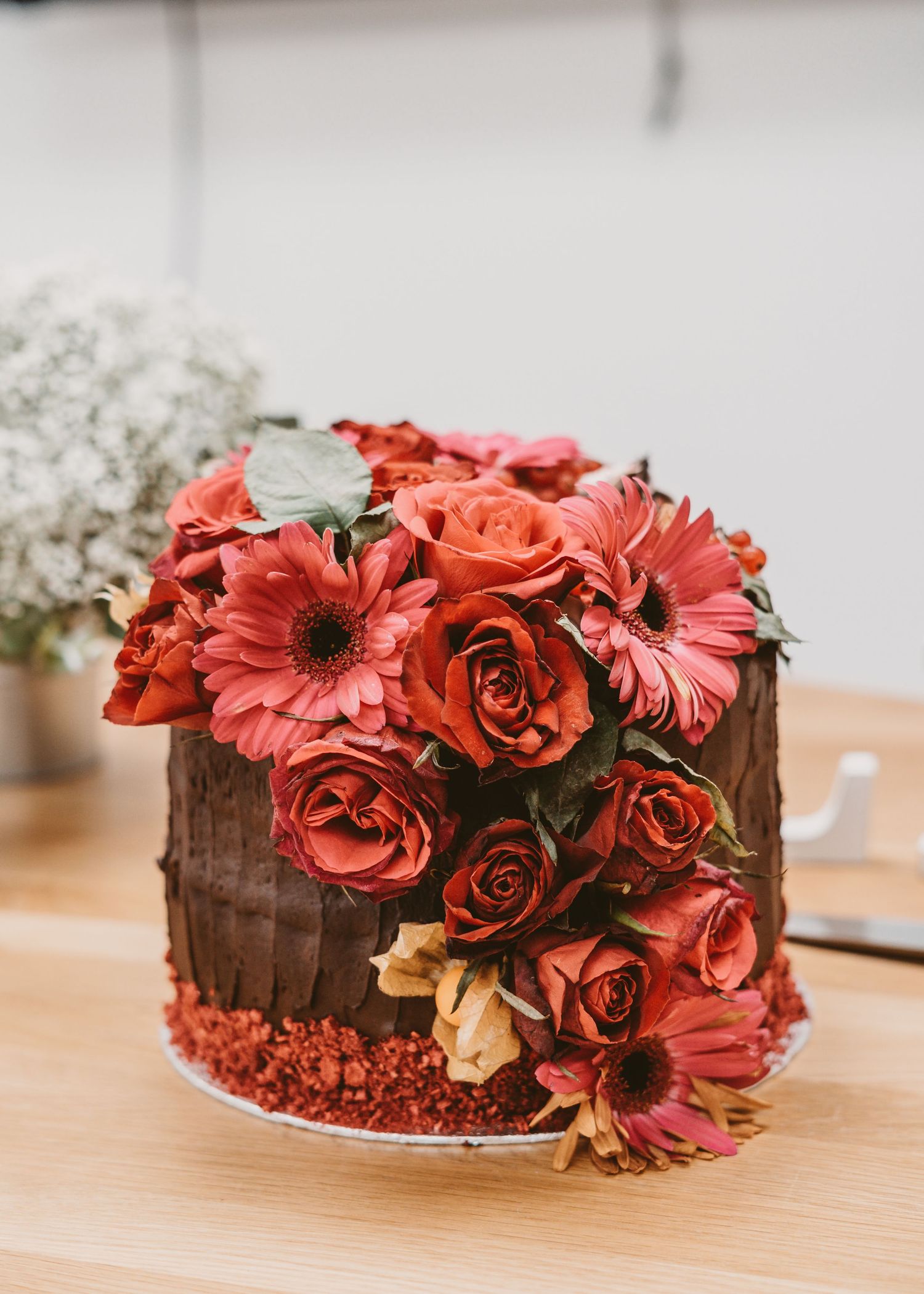 A chocolate cake decorated with coral-colored roses and gerbera daisies on a wooden table.