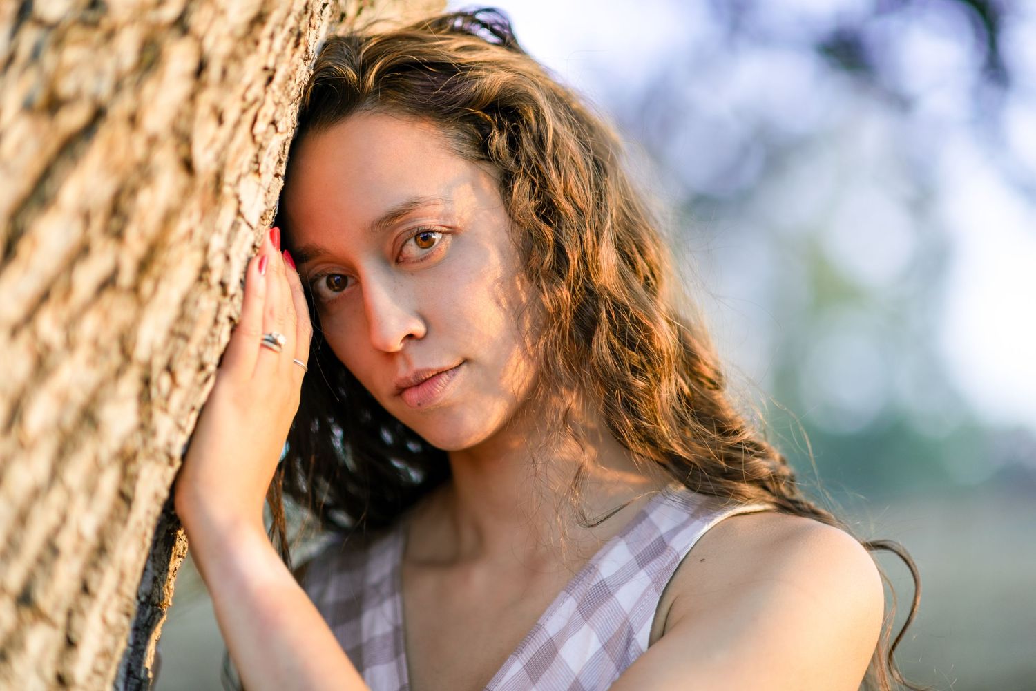 model leaning head against tree hand on tree looking at camera. long wavy brown hair with brown eyes
