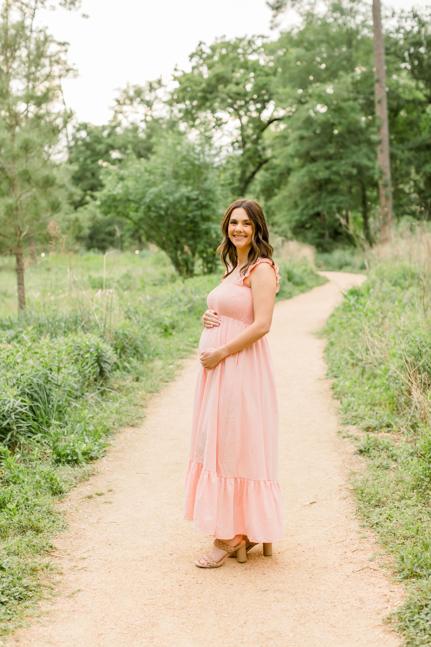 A woman in a flowing pink maxi dress poses outdoors on a nature trail.