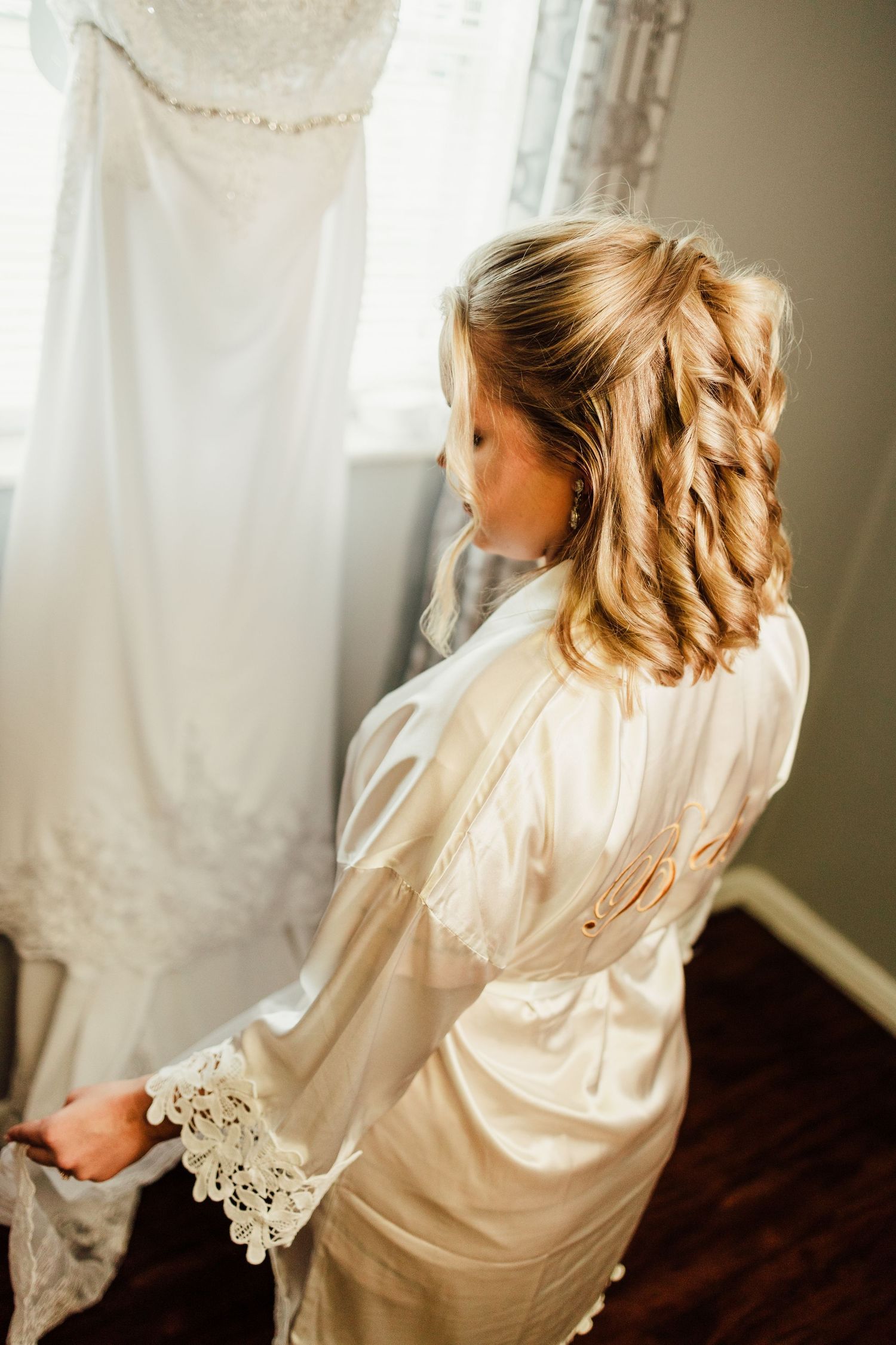Person in a silk robe looking at a hanging wedding dress in soft natural light.