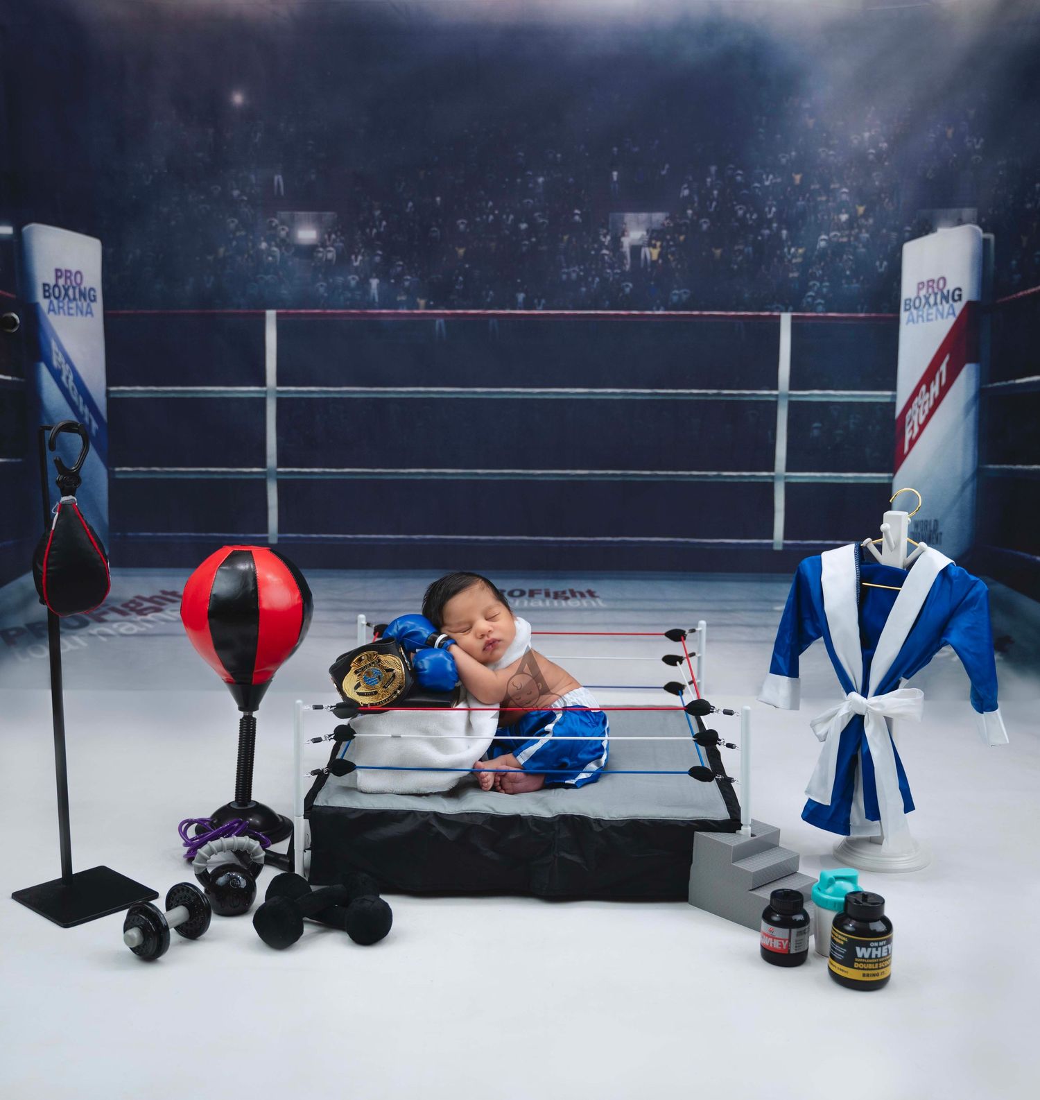 Newborn baby sleeping in miniature boxing ring with gloves, championship belt, robe, and gym equipment props.