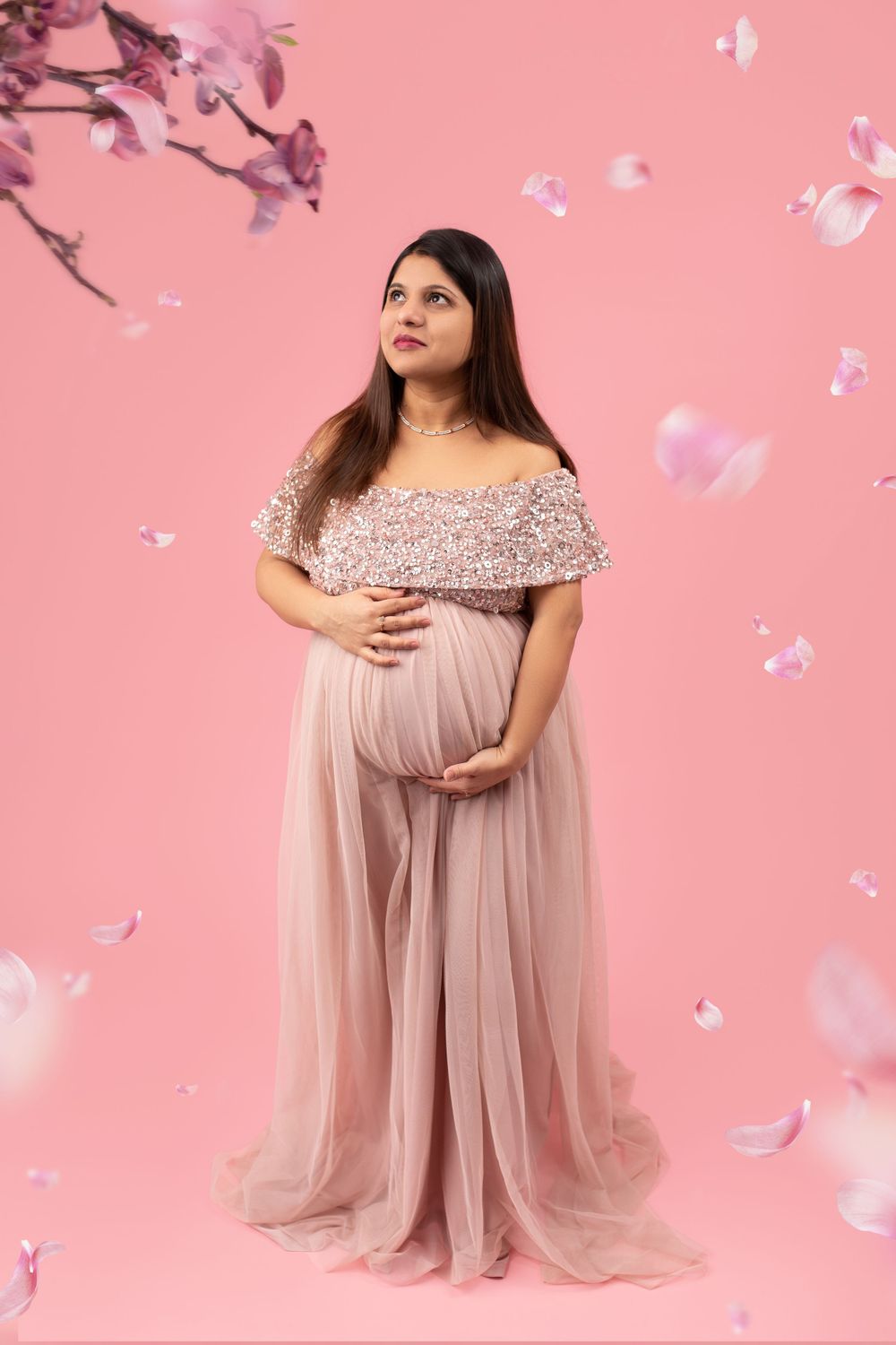 Expecting mother in flowy pink off-shoulder gown poses against pink background with falling petals.