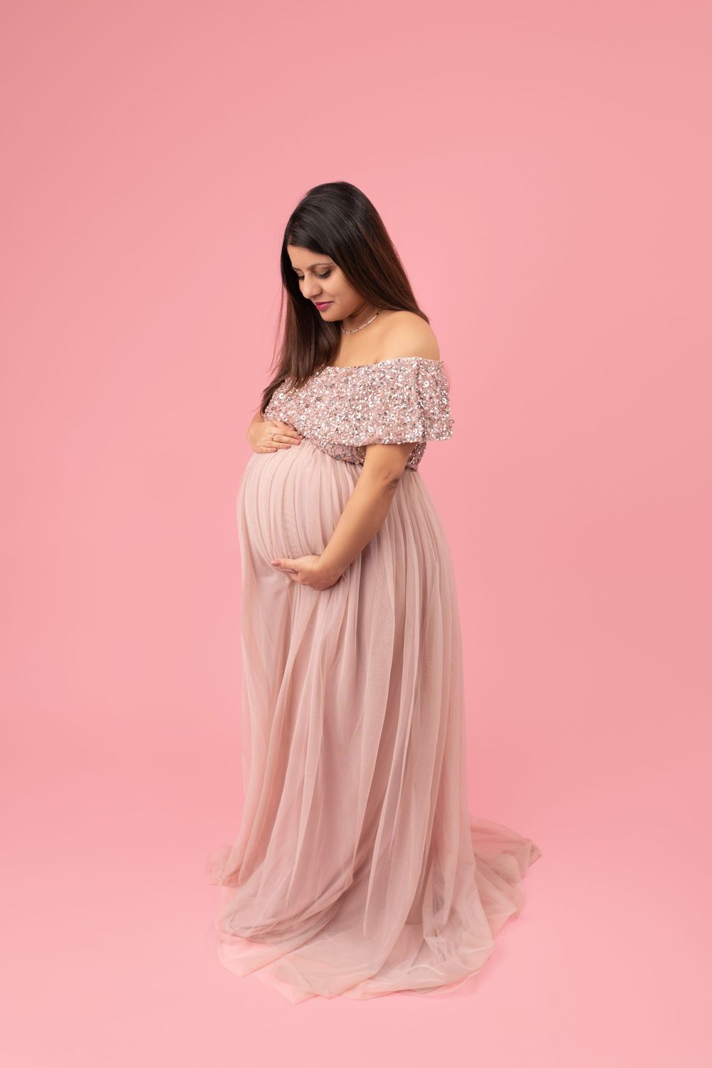 Side view of expectant mother in blush pink sequined gown against pink backdrop.