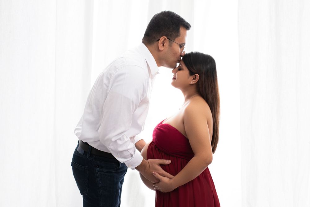 Intimate maternity portrait of couple in red dress and white shirt against white backdrop.