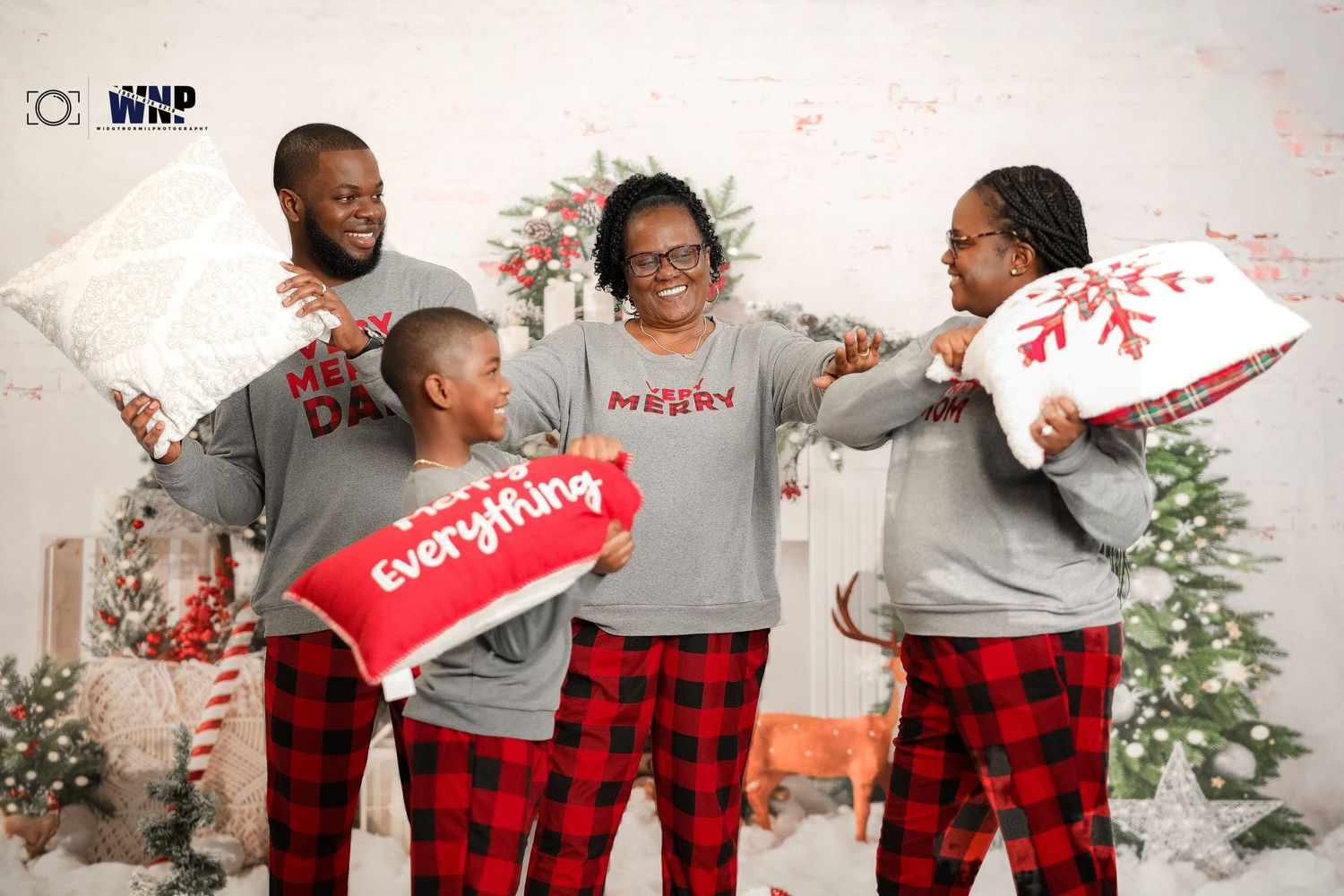 Family of four in matching plaid pajamas playfully hitting each other with pillows at Christmas.