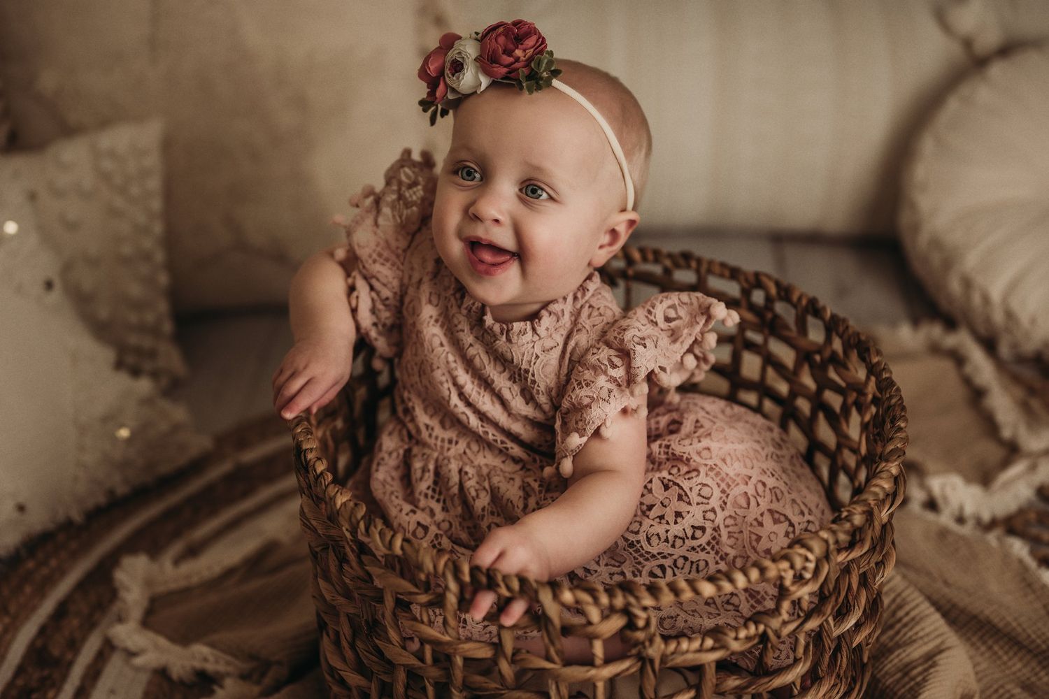 Baby sits in woven basket wearing dusty rose lace dress during vintage style photoshoot.