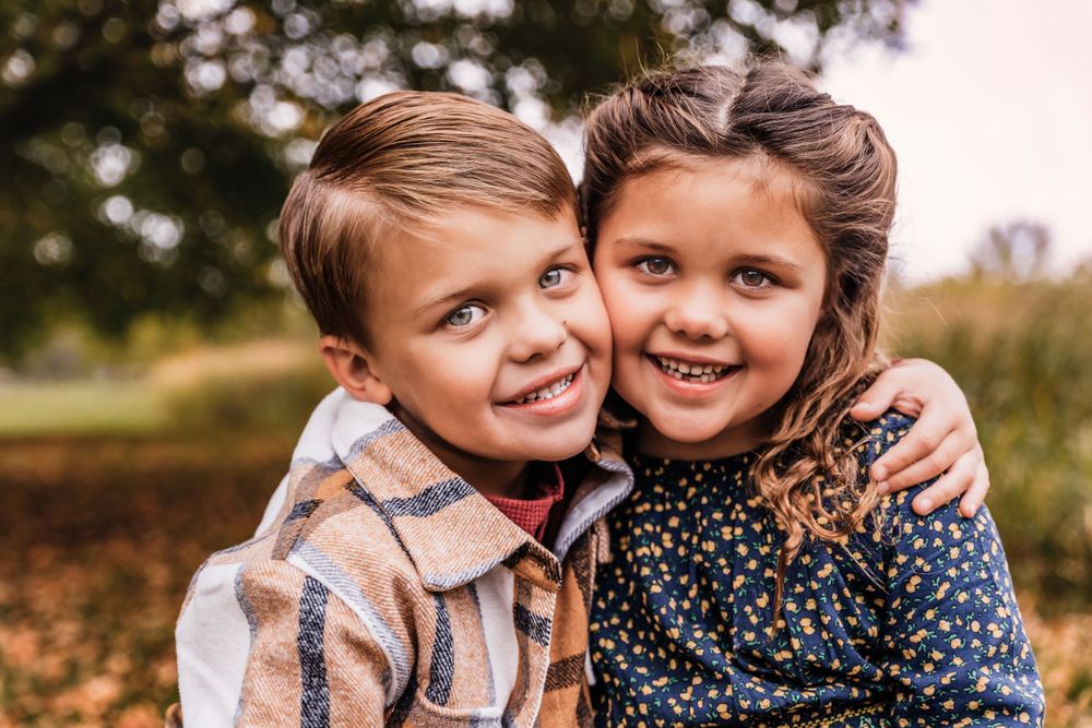 Two children embrace and smile warmly while sitting together in an autumn setting.