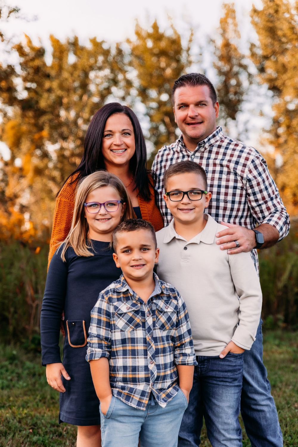 Family group portrait in casual attire against autumn trees at sunset.