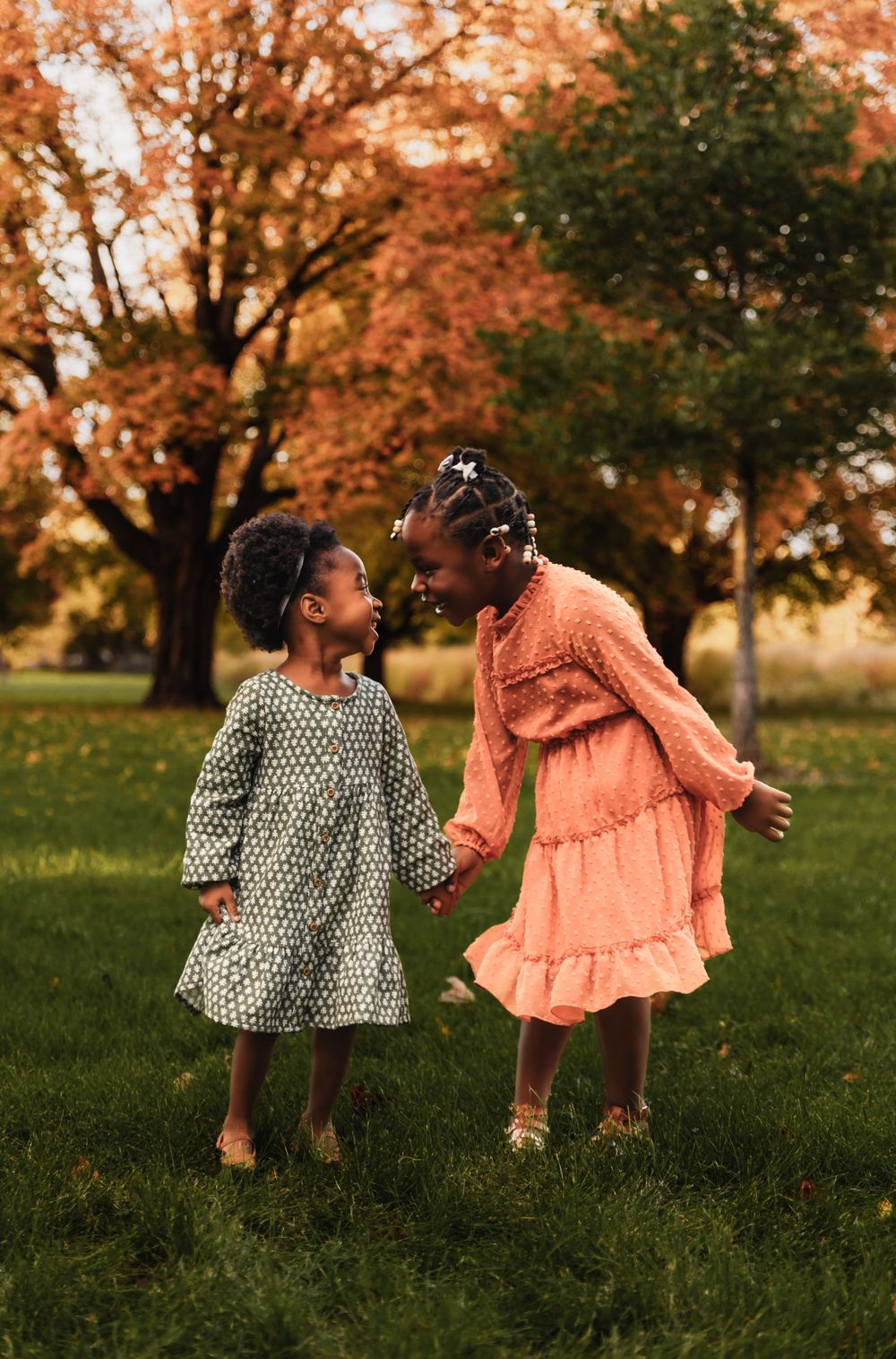 Two children in patterned dresses laugh and play together under colorful fall foliage.