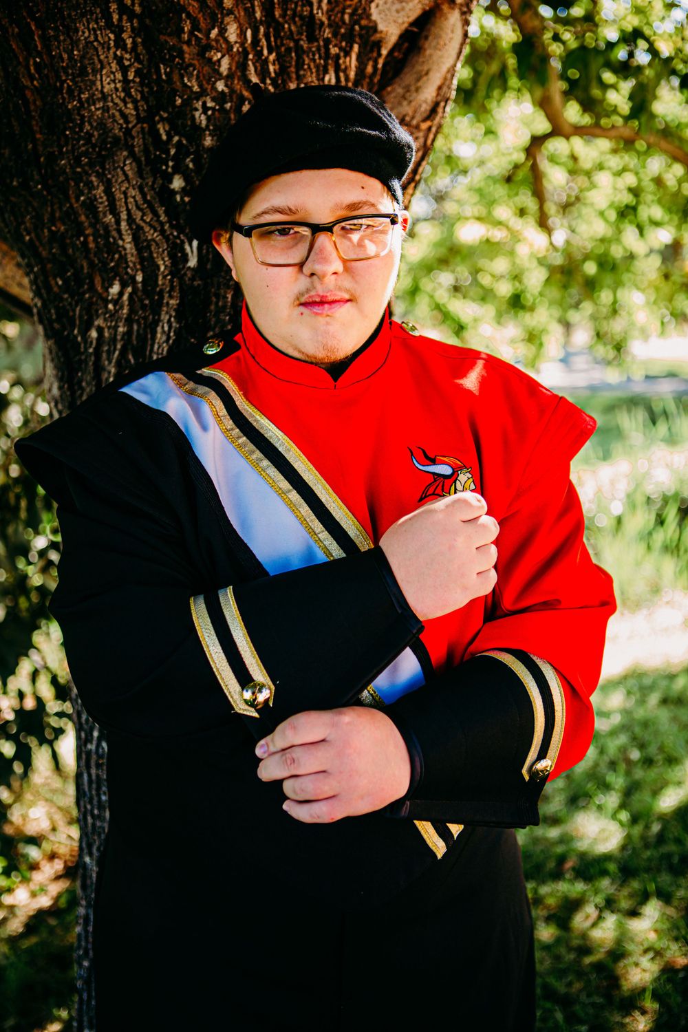 Person wearing formal red and black band uniform with gold accents poses against tree.