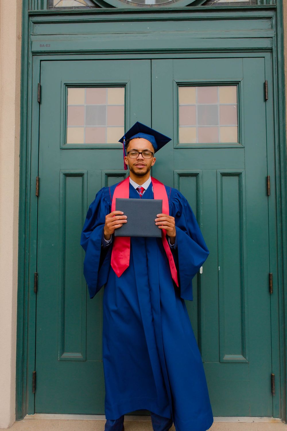 Graduate in blue cap and gown standing in front of teal doors holding diploma.