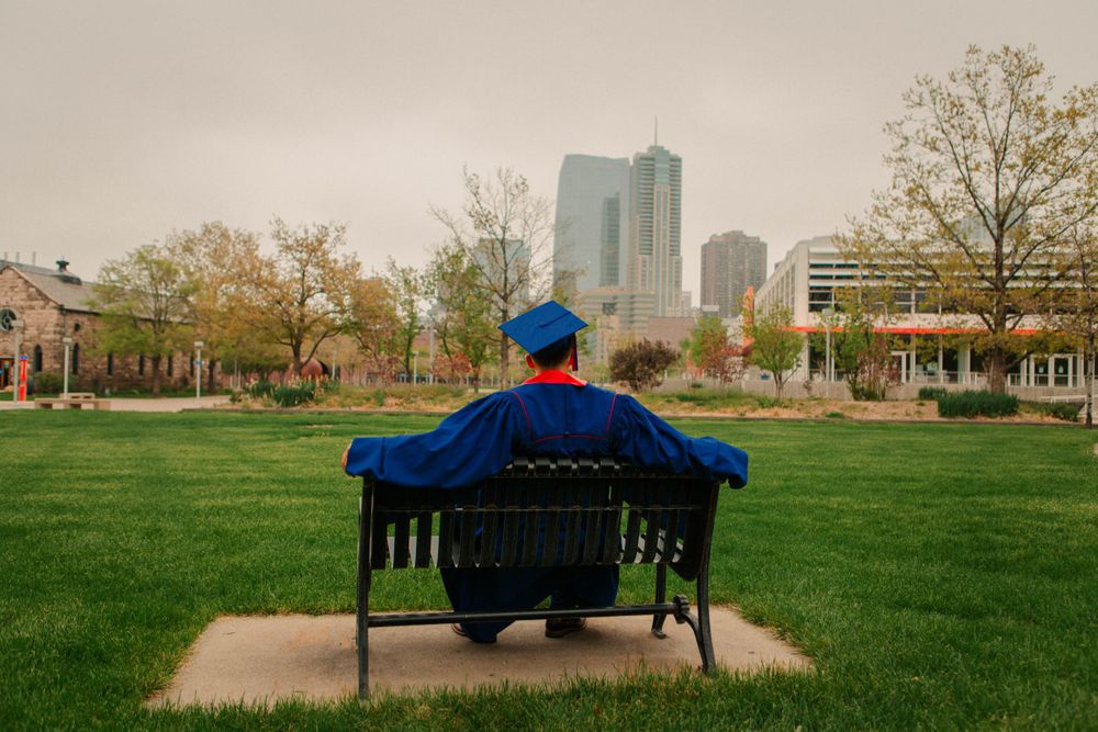 A person in blue sits alone on a bench overlooking a green lawn with city buildings in the background.
