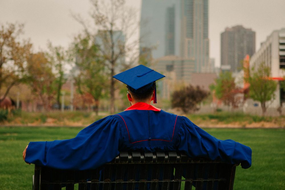 Graduate in cap and gown sitting on bench overlooking city skyline.