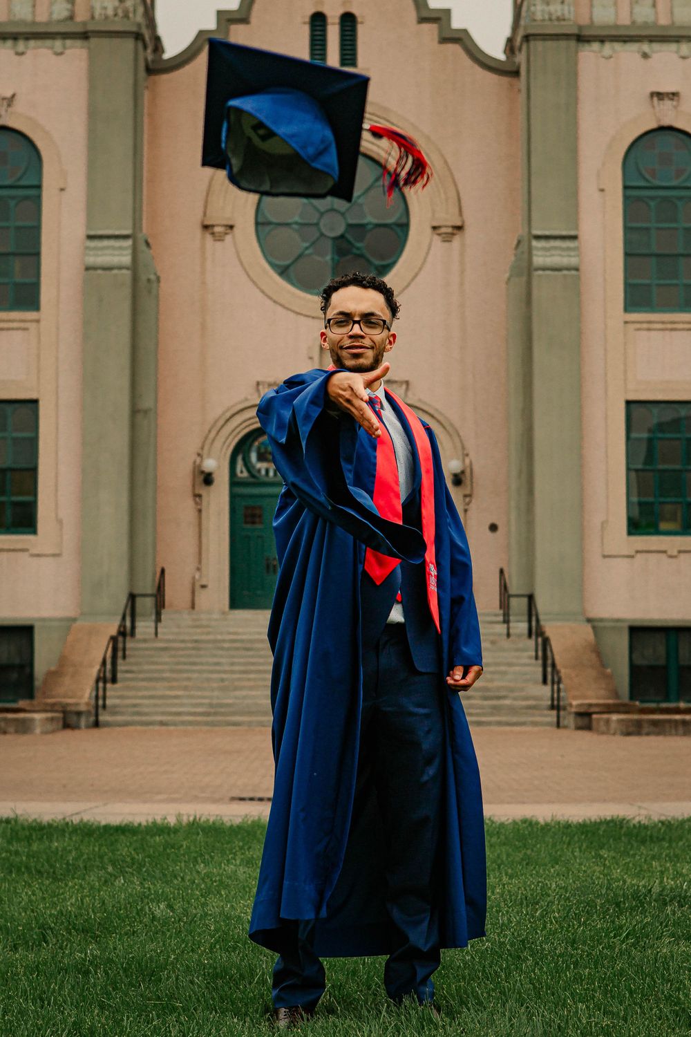 Graduate in blue cap and gown tossing graduation cap in front of historic building entrance.