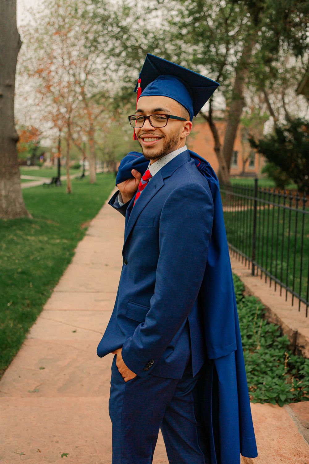 Graduate in navy suit and cap standing on campus pathway with trees in background.