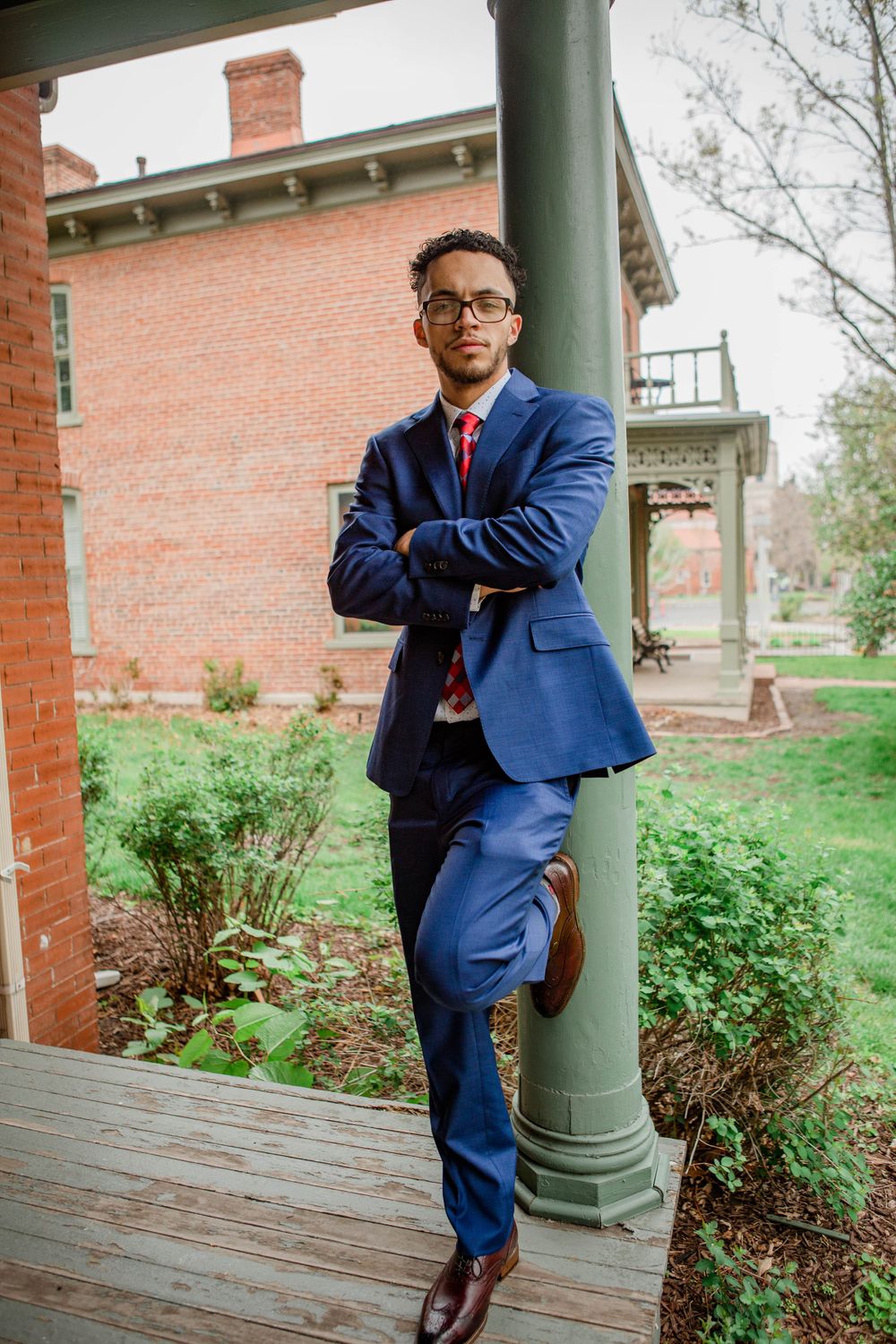 Graduate in blue suit poses confidently against brick column on campus grounds.