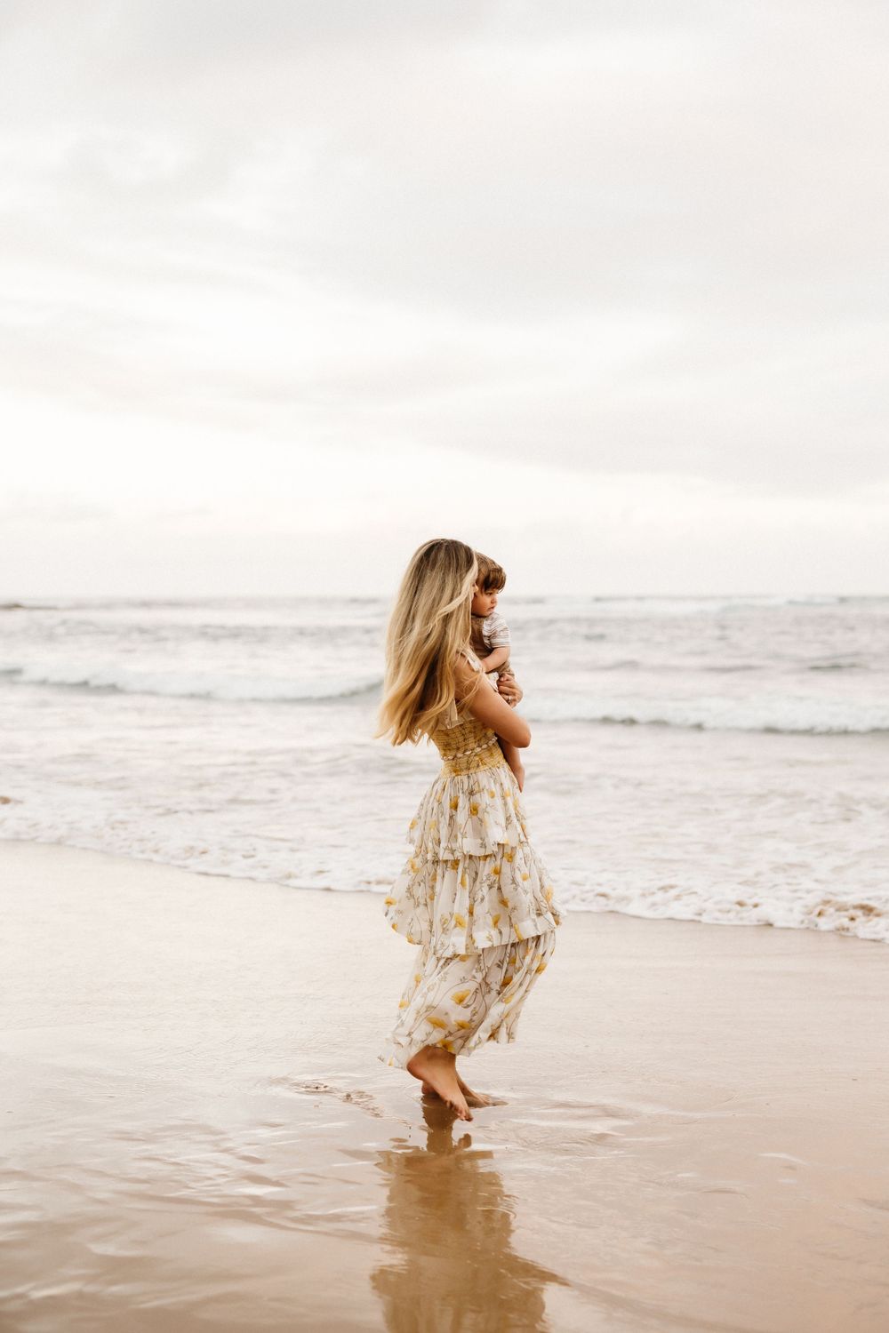 A mother and child in a flowy cream dress dances joyfully along the beach shoreline at sunset.