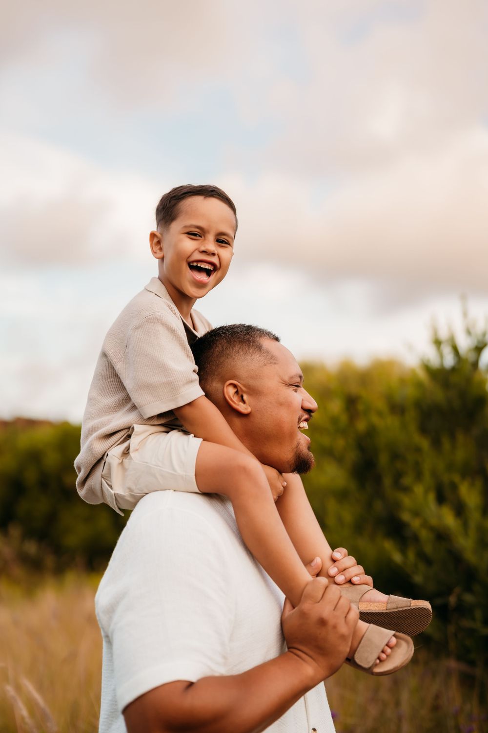 A joyful moment captured outdoors as a child rides on shoulders while both laugh together in a sunlit field.