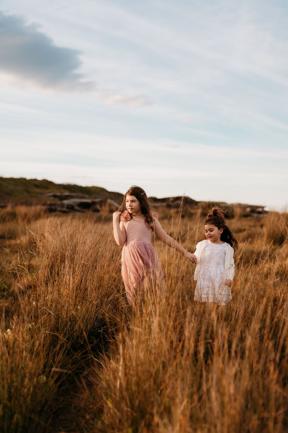 Two people in flowy dresses stand among tall golden grass in a dreamy outdoor setting at sunset.