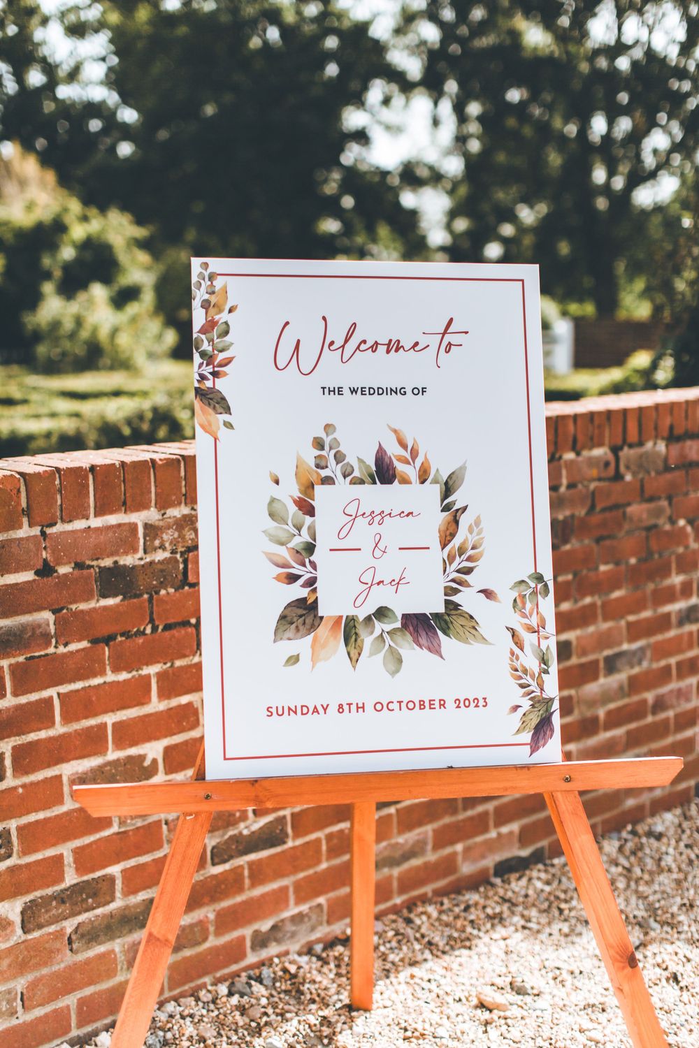 Wedding welcome sign with floral design displayed on orange easel against brick wall.