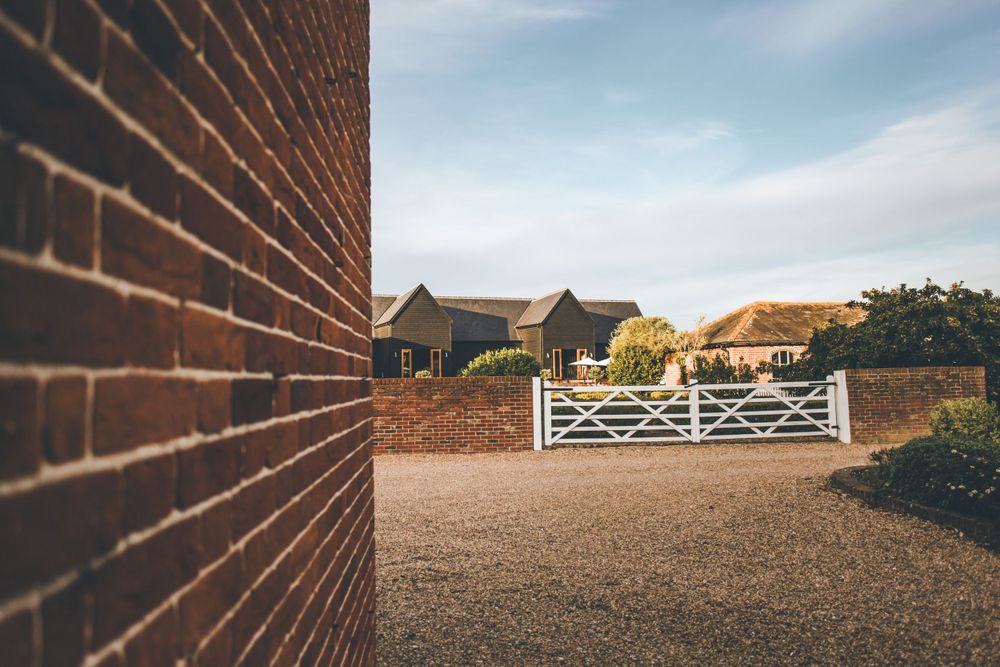 Rustic brick wall and white gate leading to countryside barn buildings.