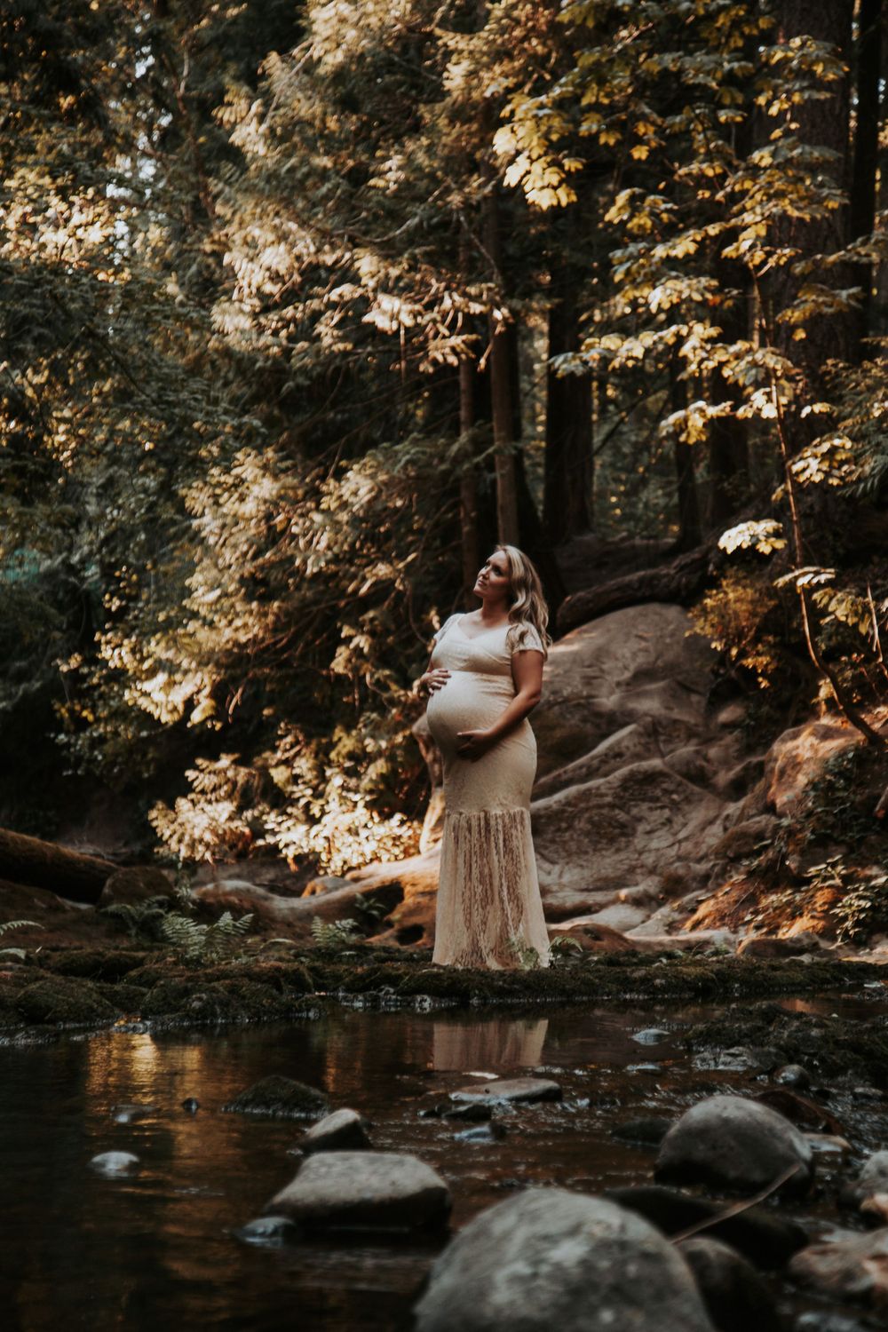Pregnant woman in a lace maternity gown gazes upward beside a woodland creek in warm golden light.