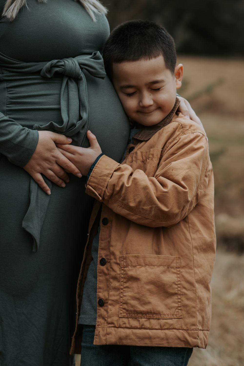Smiling boy in a tan jacket hugs his pregnant mother's belly with his eyes closed in an outdoor maternity portrait.