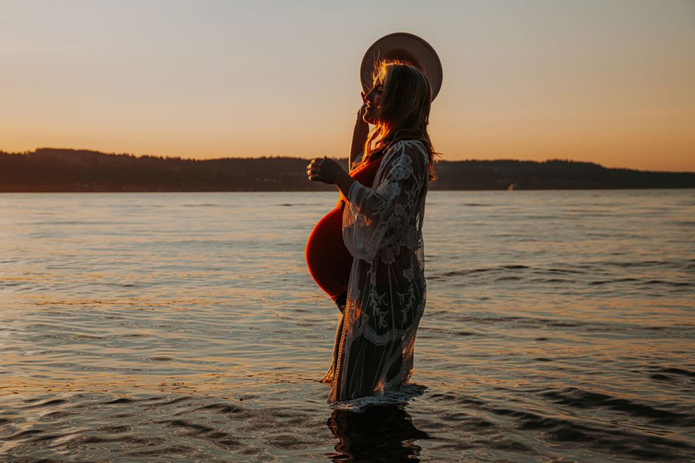 Side silhouette of a pregnant woman in a hat and lace robe standing in shallow water against an orange sunset sky.
