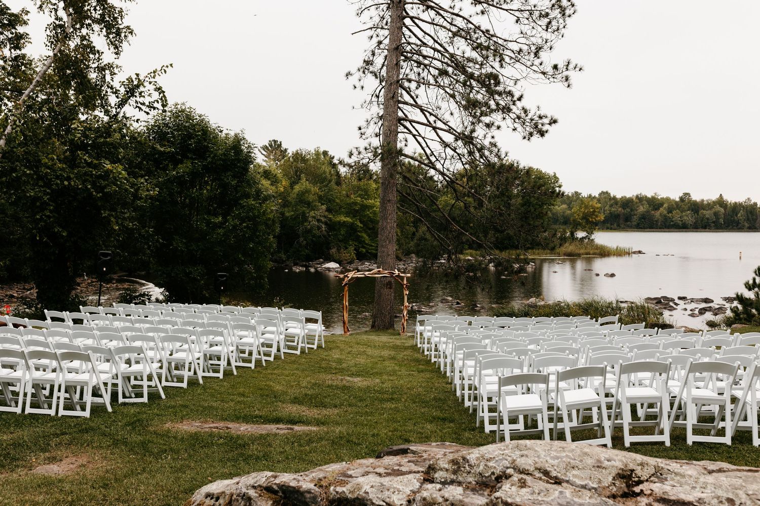 Lakeside wedding ceremony setup with white chairs and wooden arch surrounded by trees and natural scenery.
