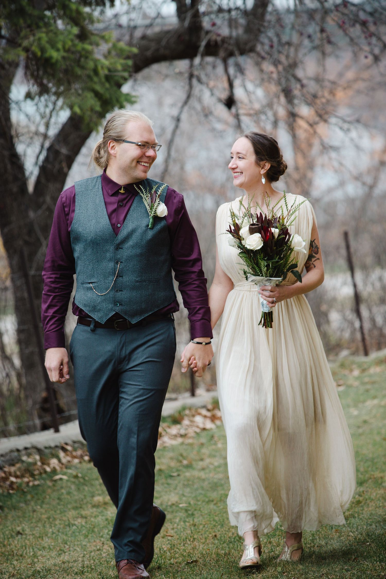 A couple walks hand in hand outdoors, with the bride holding a white floral bouquet.