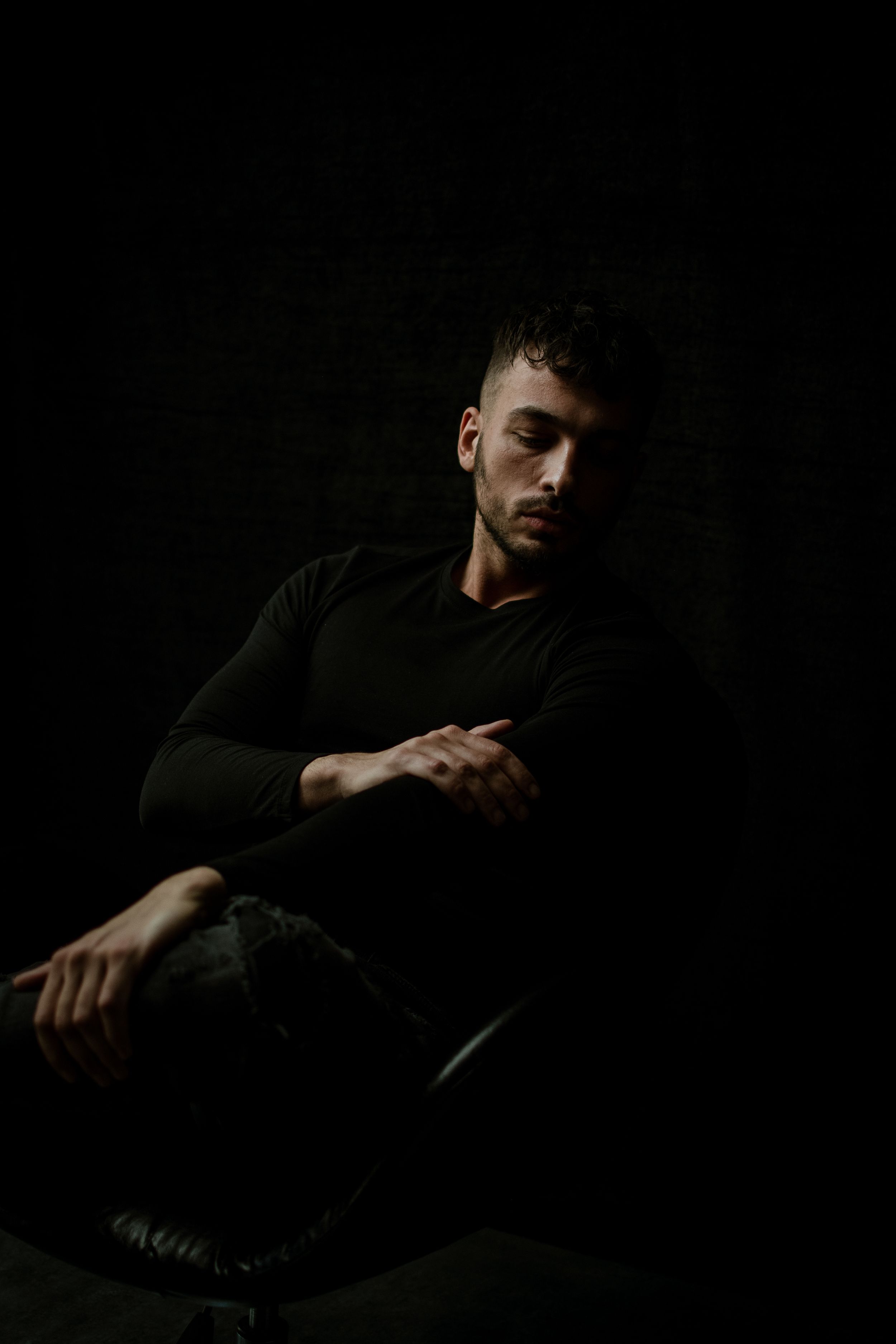 Low-key dramatic portrait of a person in dark clothing sitting in shadows.