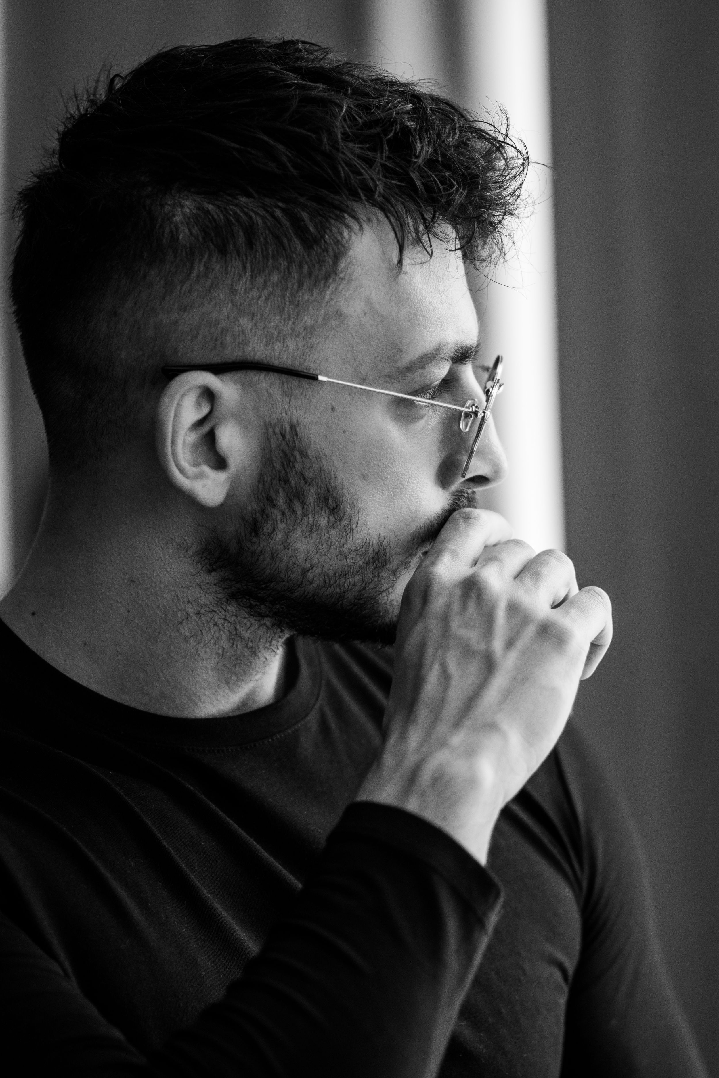 Profile view of a person with glasses in thoughtful pose against window light.