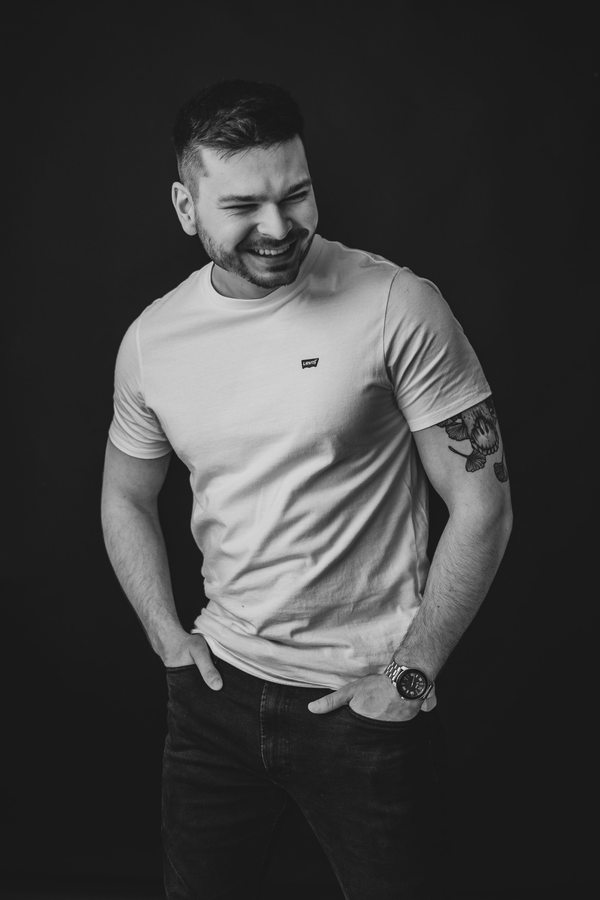 Black and white portrait of someone laughing while wearing a white t-shirt and showing arm tattoos.