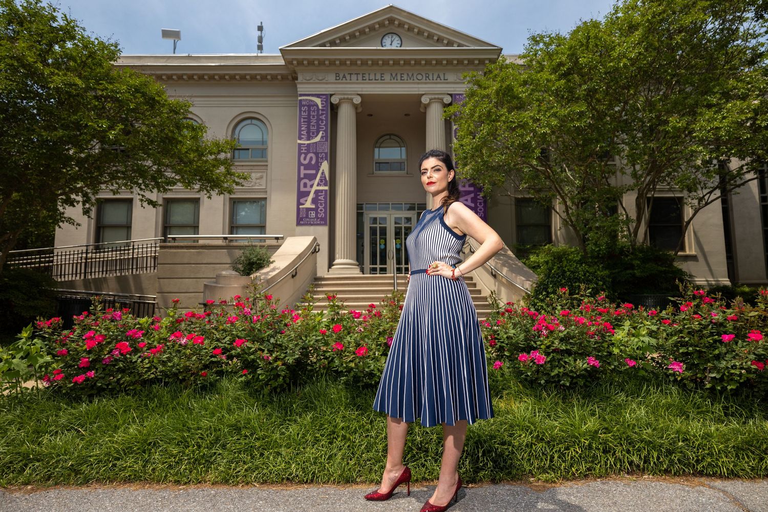 Graduate in striped dress poses confidently in front of academic building with blooming flowers.
