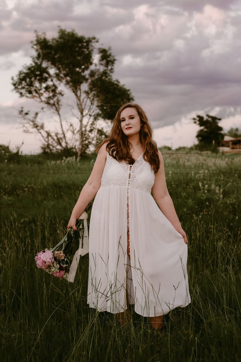 A white buttoned sundress flows in the tall grass as pink flowers are held against a cloudy sky.