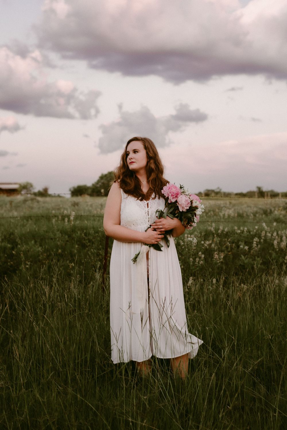 Person in white dress holds pink flowers in meadow under cloudy sunset sky.