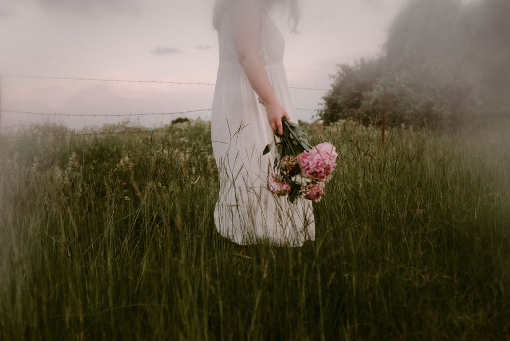 Cropped view of white dress and pink flowers against tall grass during sunset.