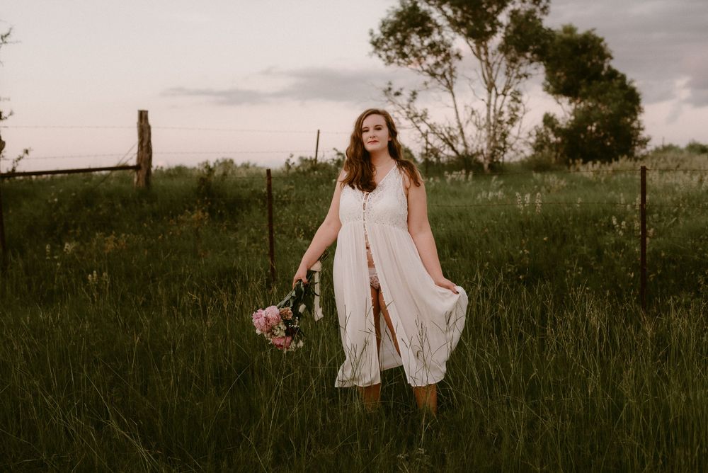 A flowing white dress catches the breeze as someone holds pink flowers in a grassy meadow at sunset.