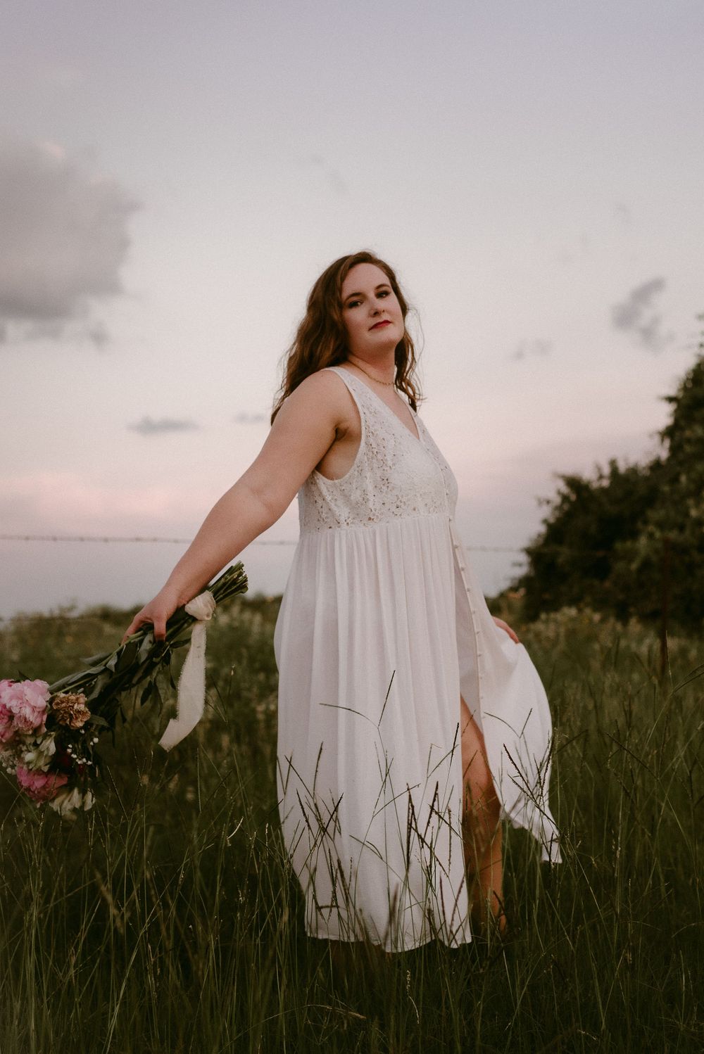 A flowing white lace dress twirls in a grassy field during golden hour while holding a pink bouquet.