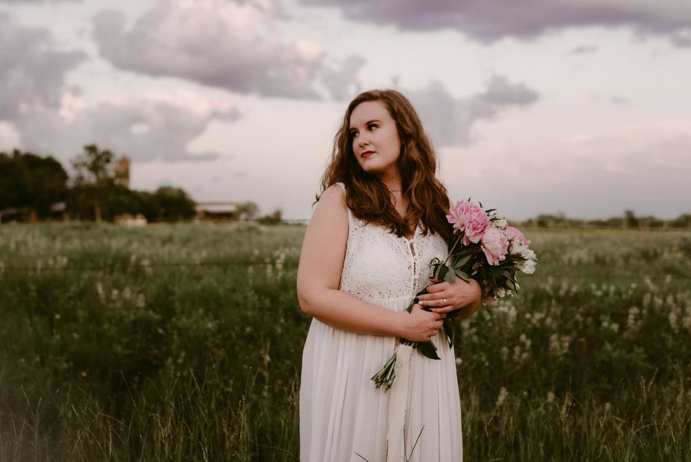 Portrait in field with white dress and pink bouquet against dramatic sky.