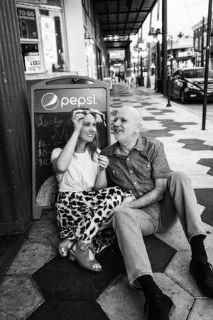 Black and white photo of couple relaxing on sidewalk near vintage Pepsi sign.