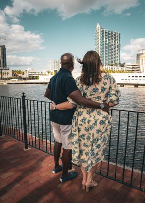 Couple overlooks waterfront from wooden deck with city skyline in background.