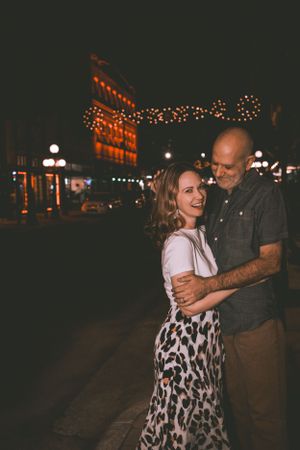 Couple shares intimate moment on city street at night illuminated by string lights.