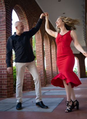 Dancing sequence showing couple in red dress and black attire moving along brick colonnade.